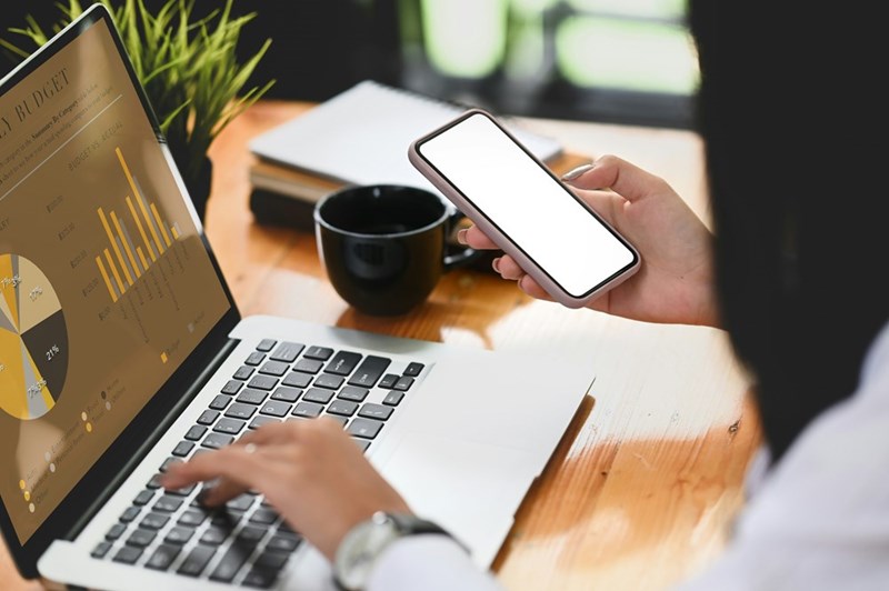 Woman wearing a shirt sitting in a desk holding a phone and looking at a graph in front of her in the computer.