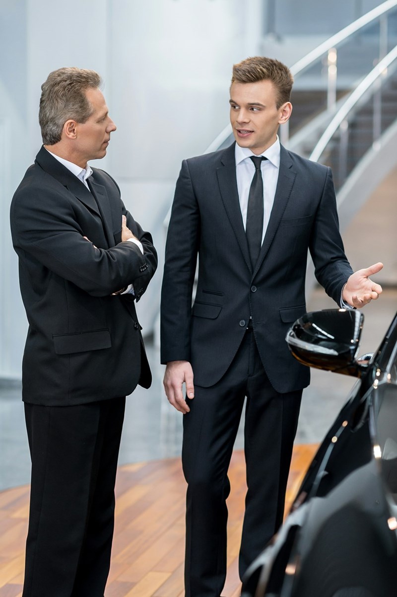A well suited sales man is showing another suited man a car.