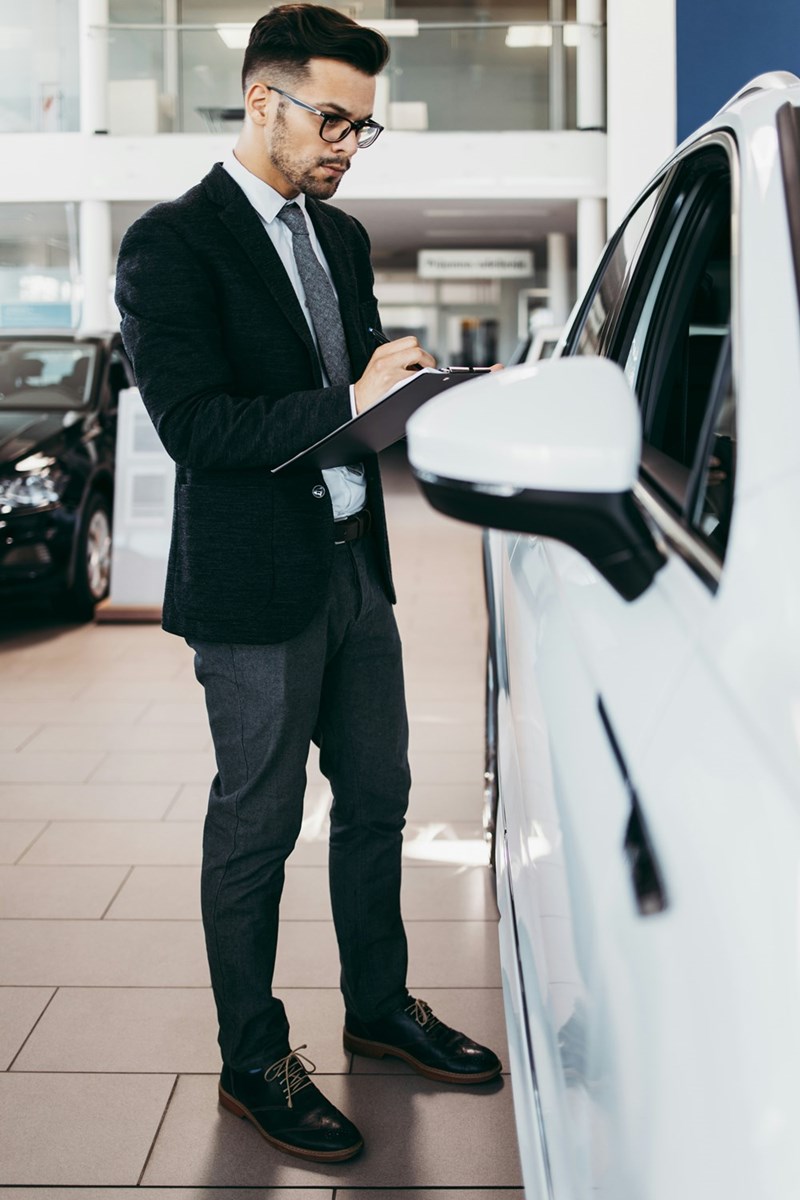 A black suited salesman holding a pen and a deal on paper while looking a a car in a car dealership.