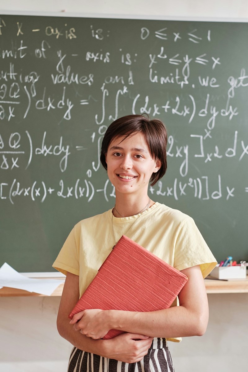Young teacher holding a notebook stands smiling in front of a classroom chalkboard filled with math equations.