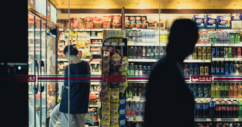 A silhouette passes in front of the glass door of a convenience store with a female shopper inside 