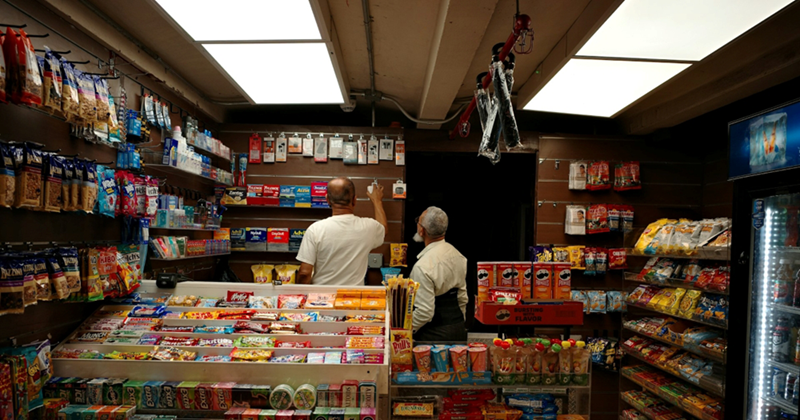 A male convenience store employee helps a customer find something on the back wall