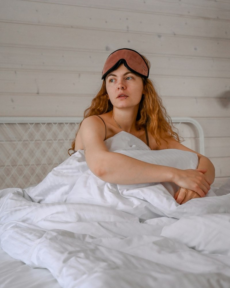 Woman sitting in bed with sleep mask and white blankets, looking tired after waking up.
