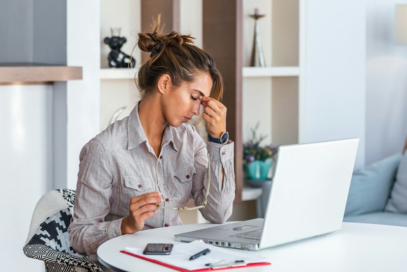 A woman looks stressed while working on her laptop at home, holding her glasses in frustration.