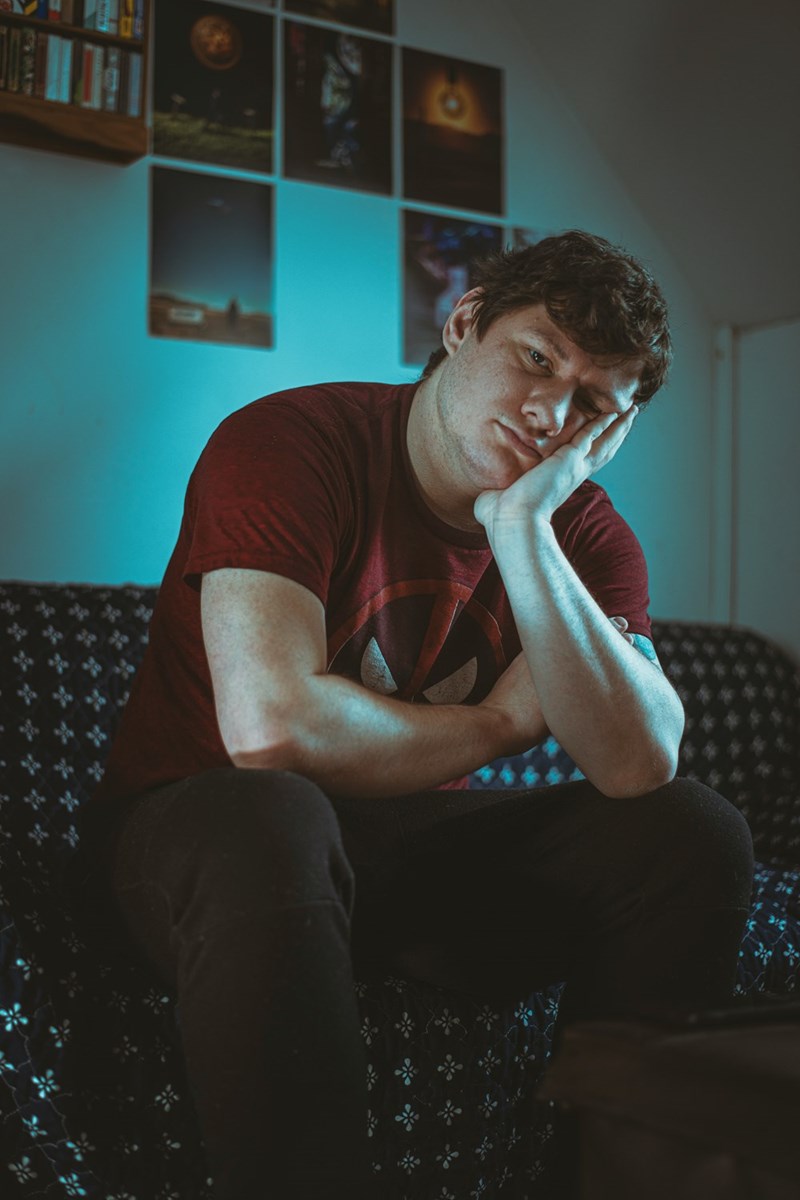 Young man sitting on couch with tired, concerned expression in dimly lit room.