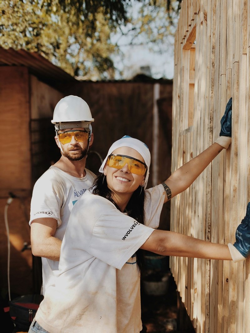 Couple wearing safety gear while working together on a wooden wall at an outdoor construction site.
