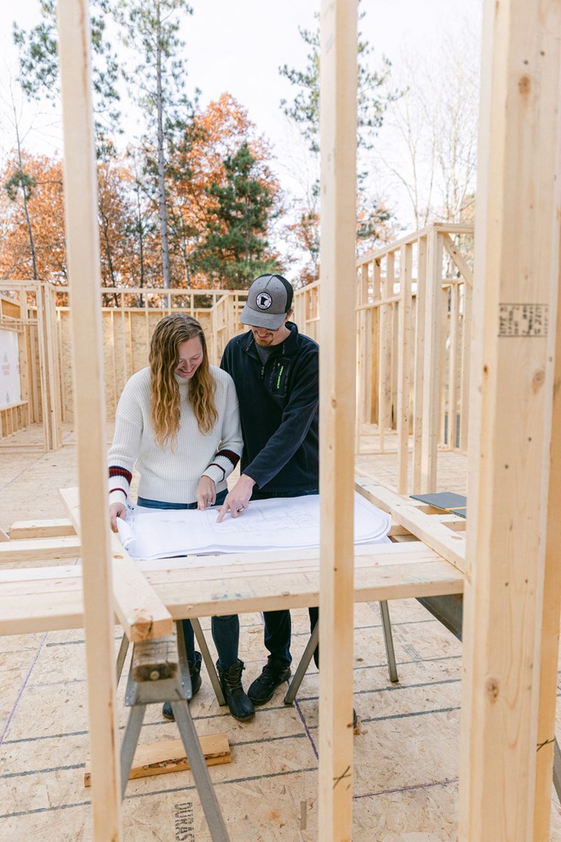 Couple reviewing house blueprints together inside a wood-framed home under construction.