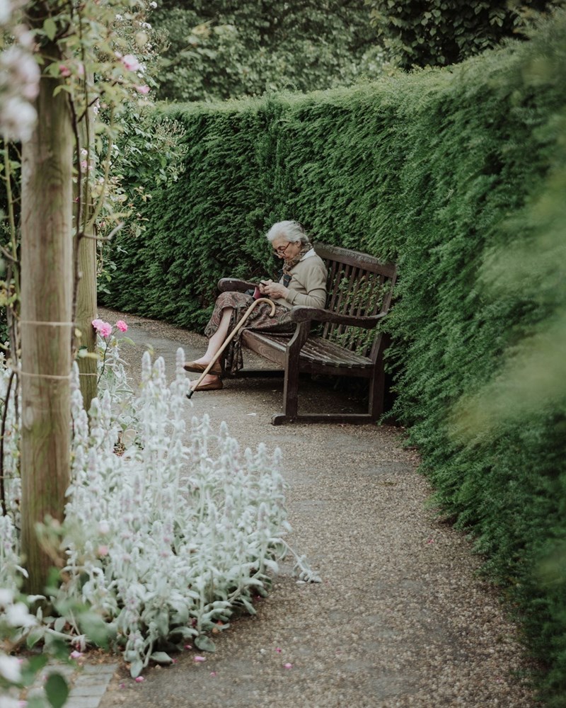 An elderly woman sitting in a bench in a side-yard filled with trees, plants, and flowers.