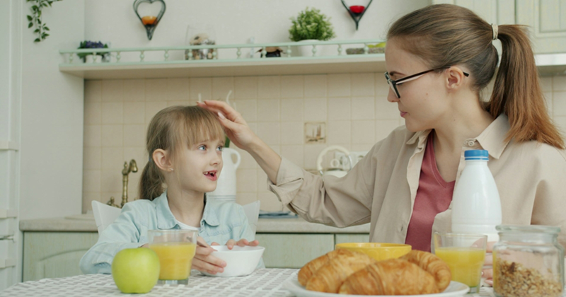 A young woman touches the hair of a little girl while they both sit at a kitchen table