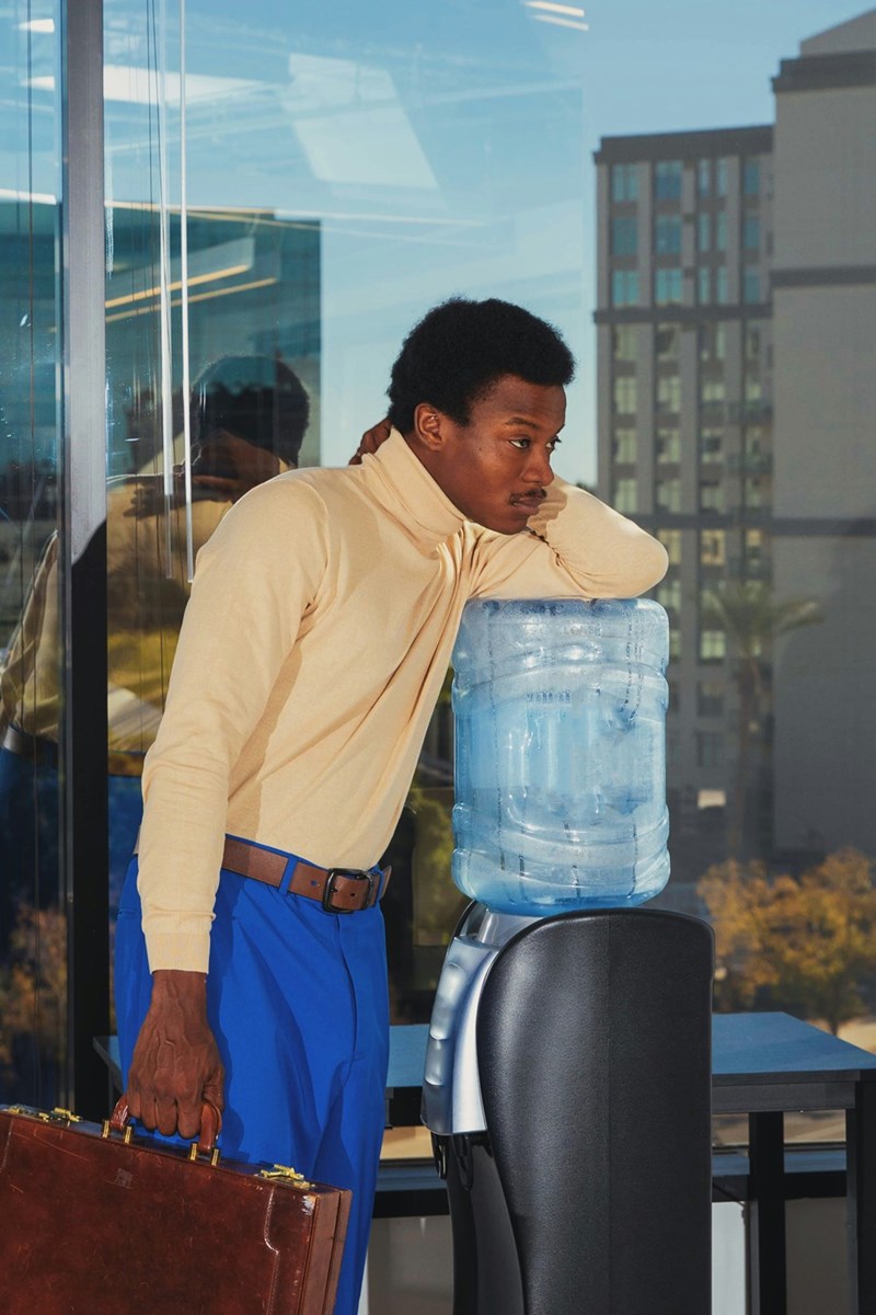 Worker holds briefcase while leaning on watercooler, looking very bored.
