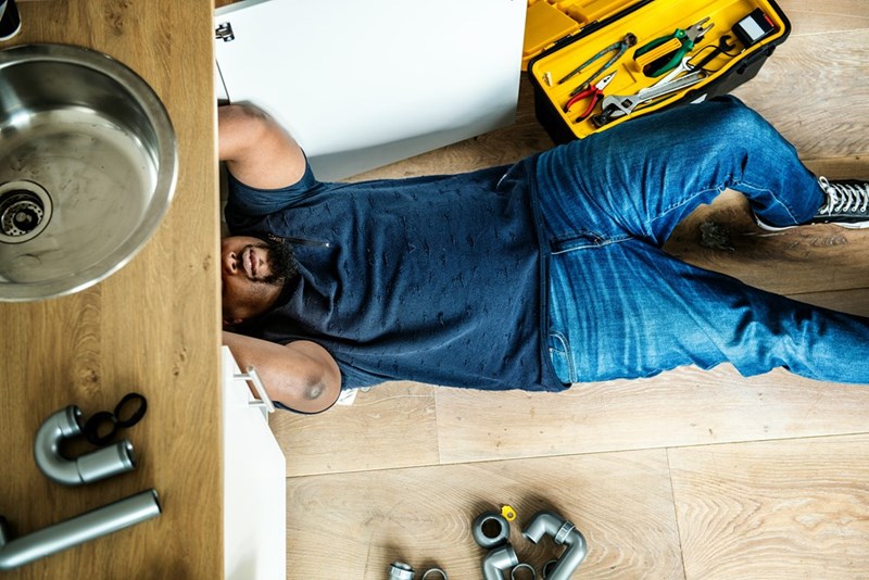 Plumber wearing jeans is laying on the floor checking the kitchen faucet.