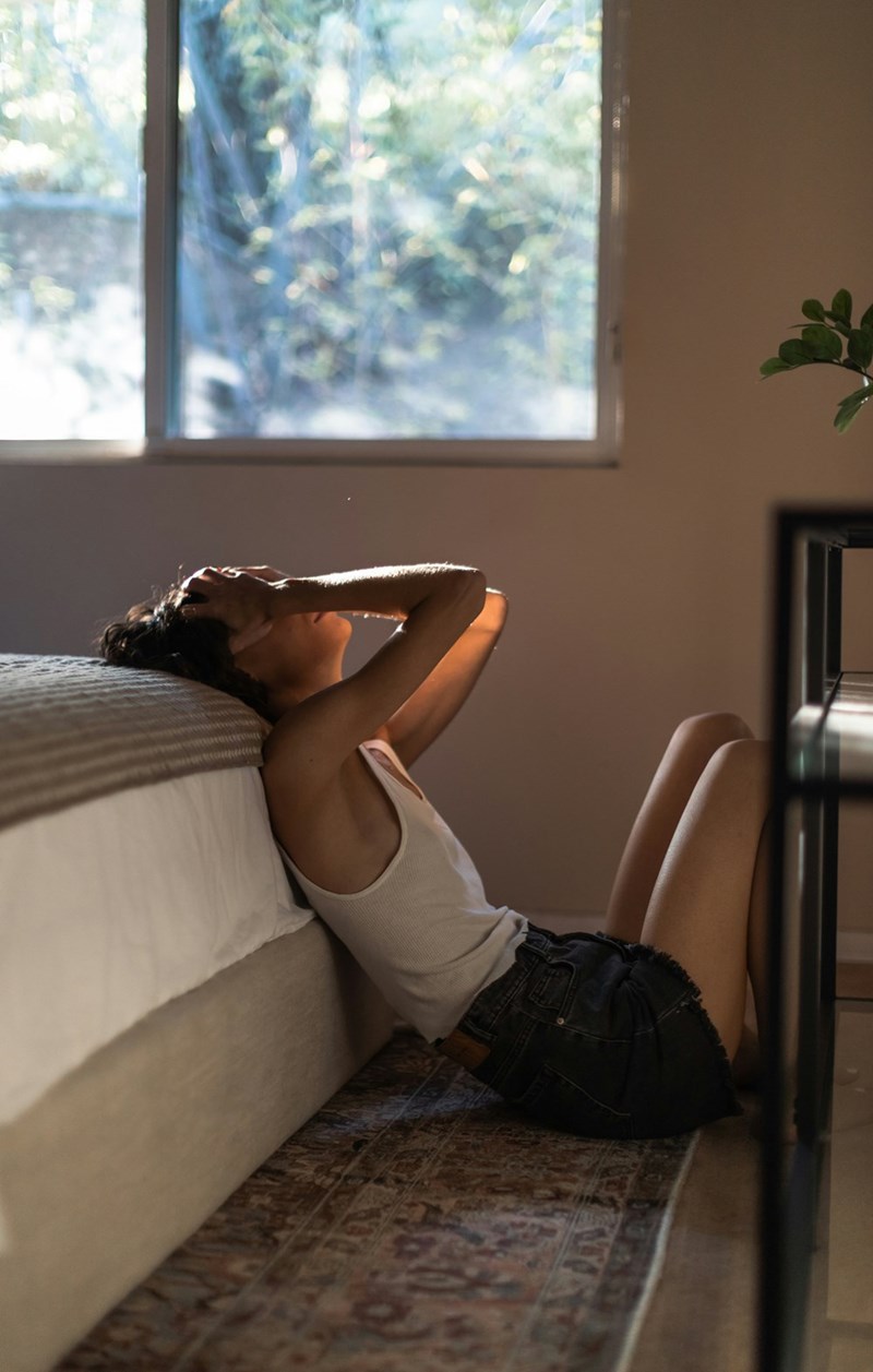 Sad woman sitting on the flor laying her back on her bed.