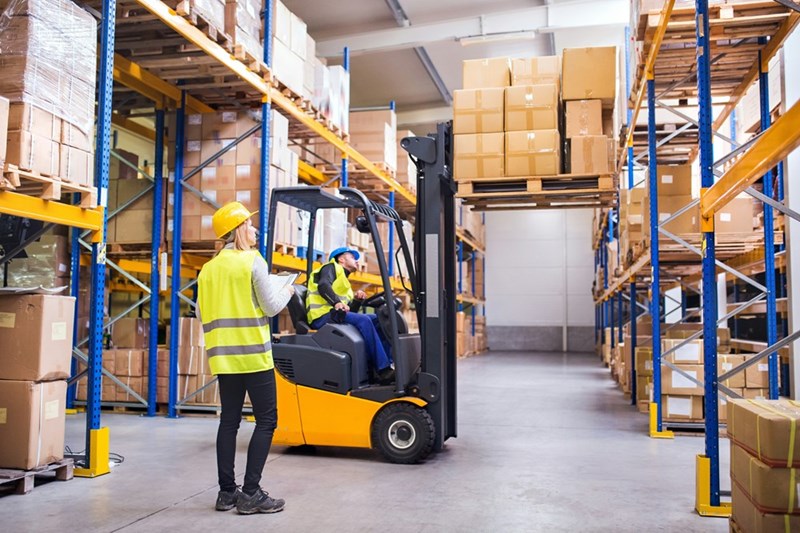A young warehouse employee works alongside a man sitting in a forklift.