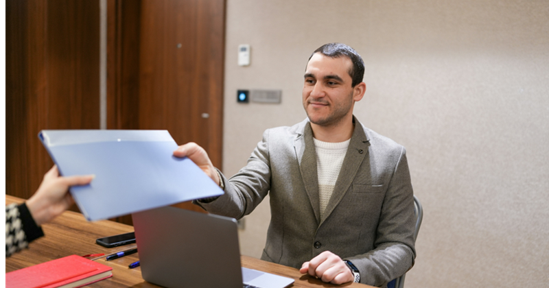 A man sits at a table wearing a jacket, handing over a portfolio.