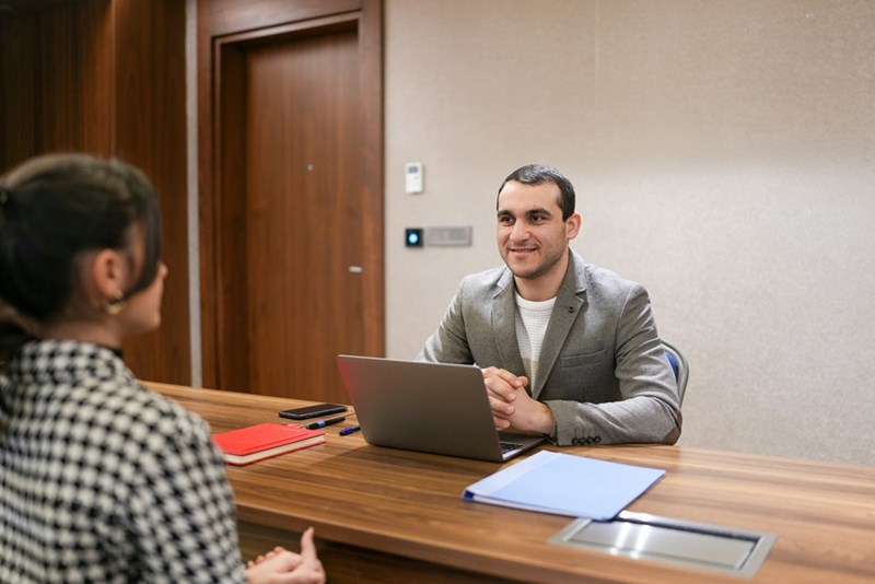 A man sits at a table wearing a jacket in front of a laptop, opposite a recruiter.