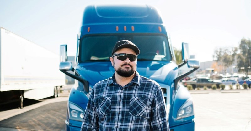 A man wearing sunglasses and a flannel shirt stands in front of his blue truck.