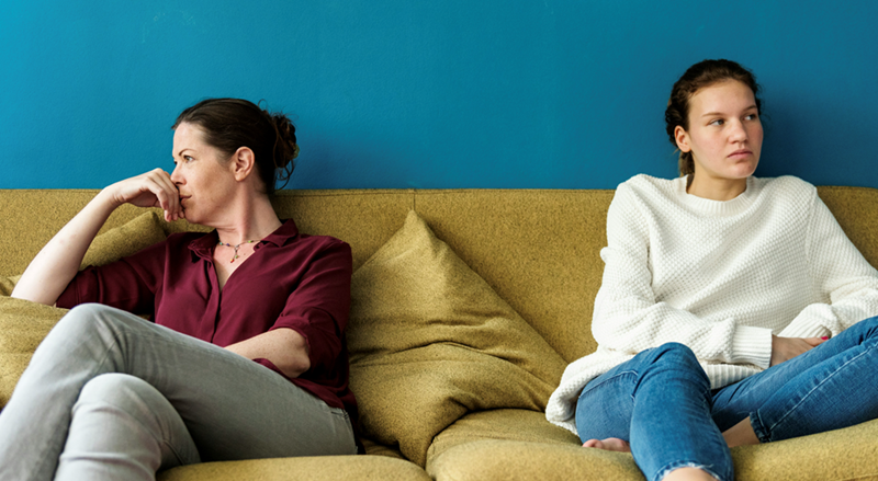 Mother and daughter avert eye contact while arguing on a sofa.