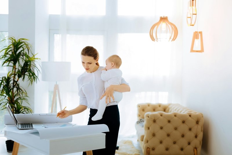 A working mom multitasks with her baby in one arm while reviewing documents at her desk.