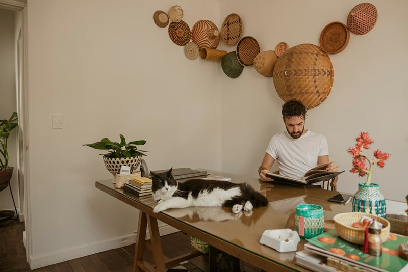 Man reading at a dining table while a black-and-white cat lounges beside him in a cozy home.