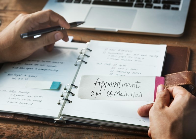 Hands holding a paper with an appointment text written on it and an agenda laying on a desk.