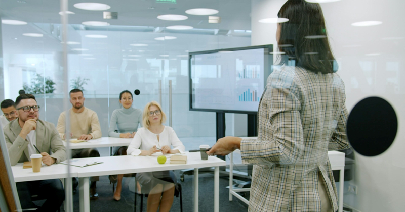 A female employee behind a glass wall presents a meeting to coworkers