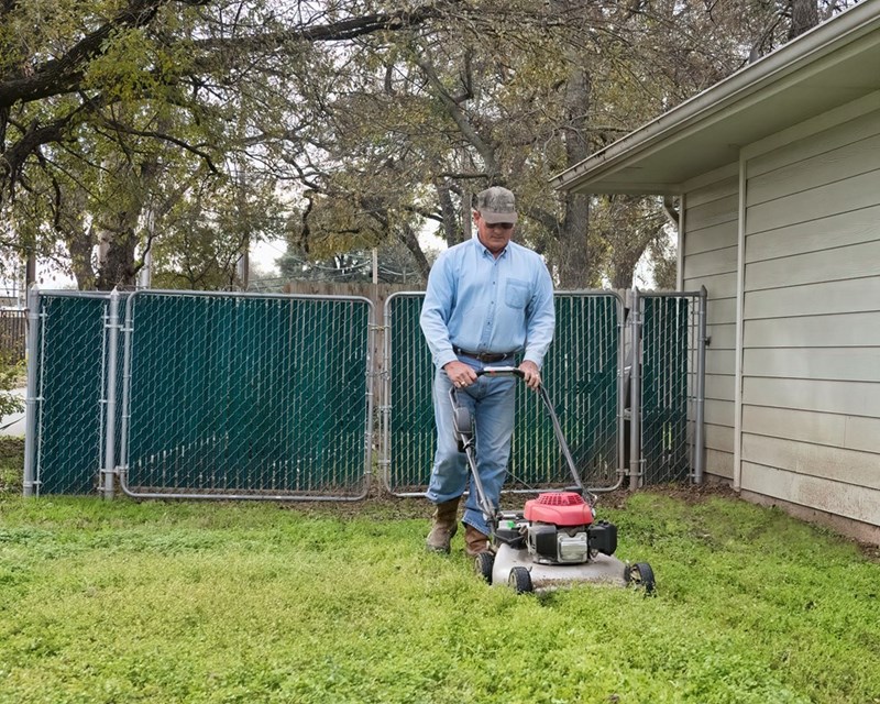 Homeowner mowing backyard lawn with push mower beside suburban house.