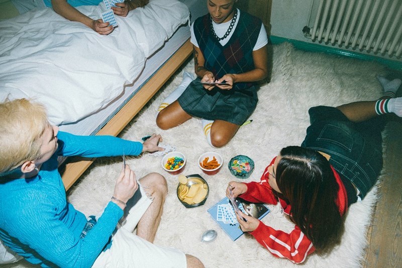 Friends playing cards on a rug with snacks during a cozy indoor game night beside a bed.