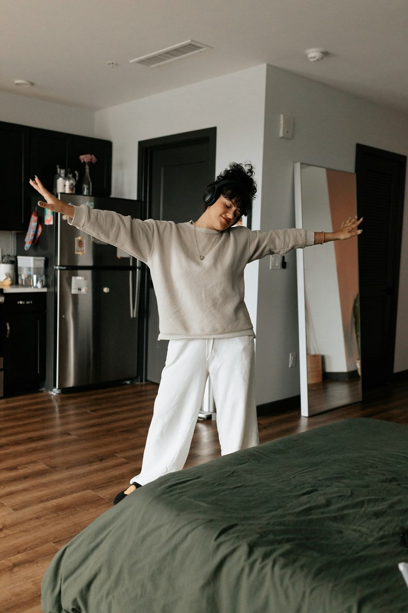 Woman in loungewear dancing with headphones in a modern apartment bedroom near the kitchen.