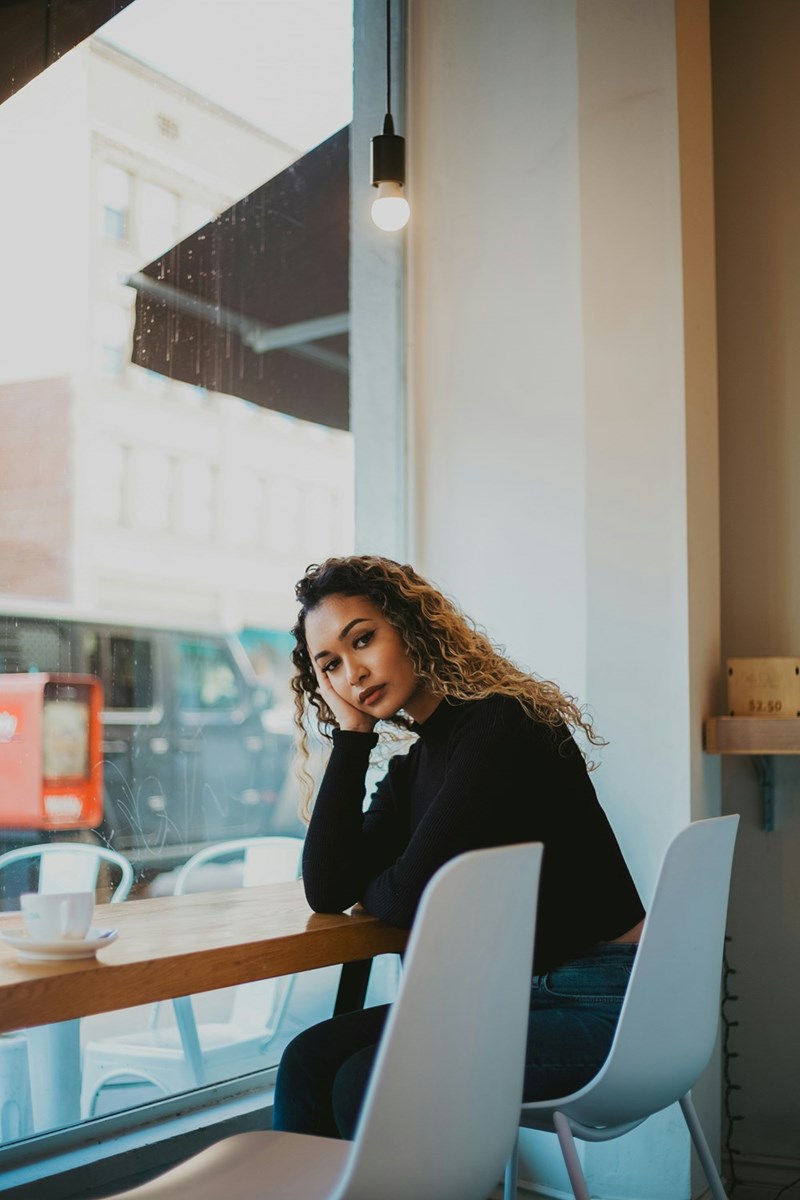 Woman sitting alone in a coffee shop beside a window, looking thoughtful during a quiet moment.