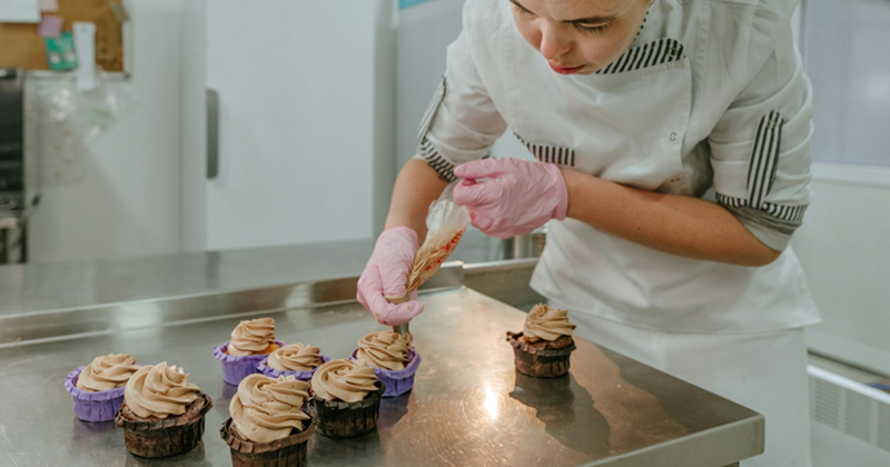 A baker ices a chocolate cupcake with a piping bag