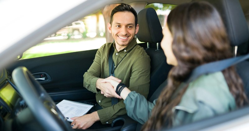 A male driving instructor shakes hands with a teenage female student while they are inside a vehicle