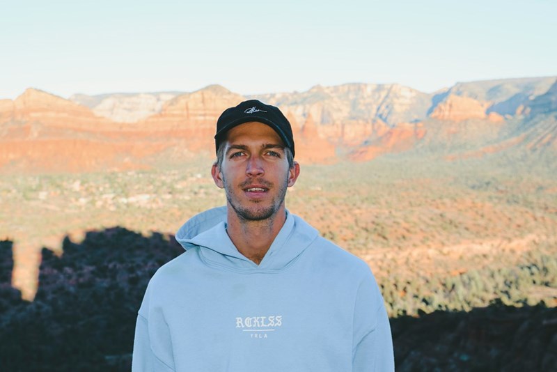 Man in hoodie standing outdoors with red rock desert mountains and scenic landscape behind him.