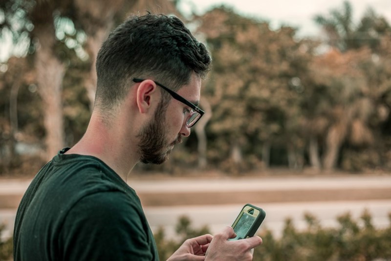 Man using smartphone outdoors, wearing glasses and looking down at screen in a park-like setting.