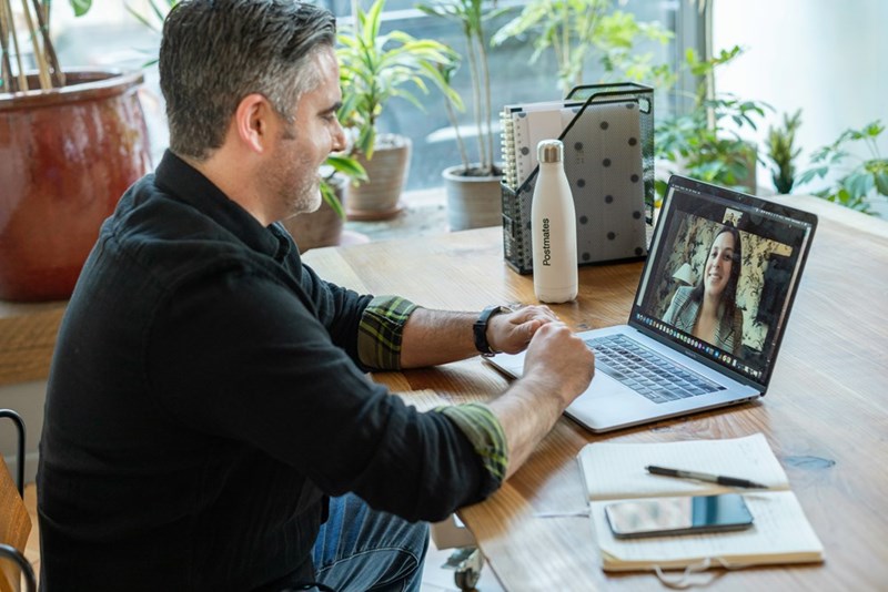 Man on laptop video call at desk with plants, smiling during remote meeting at home office.