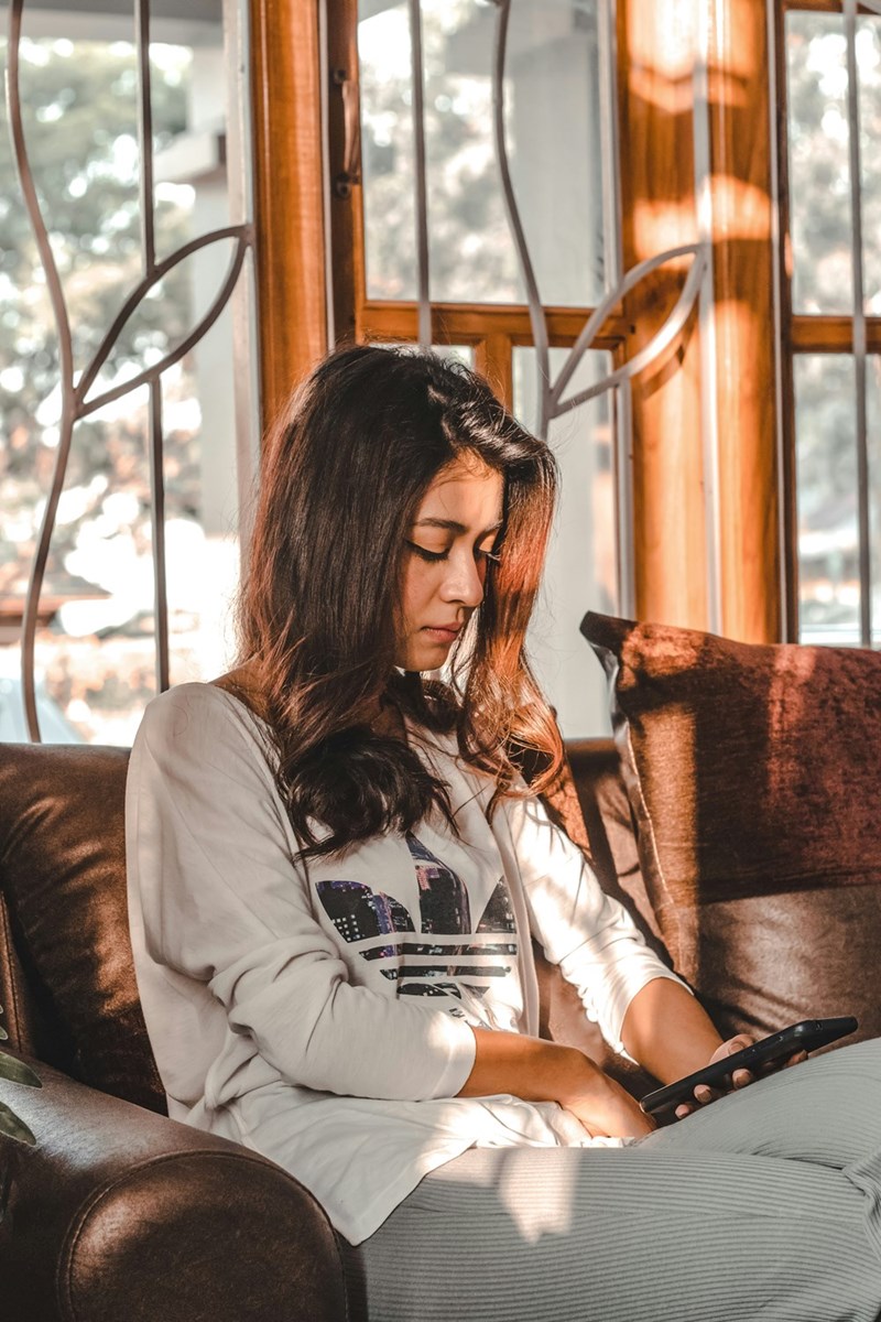 Woman using smartphone on couch by window, bathed in warm sunlight at home.