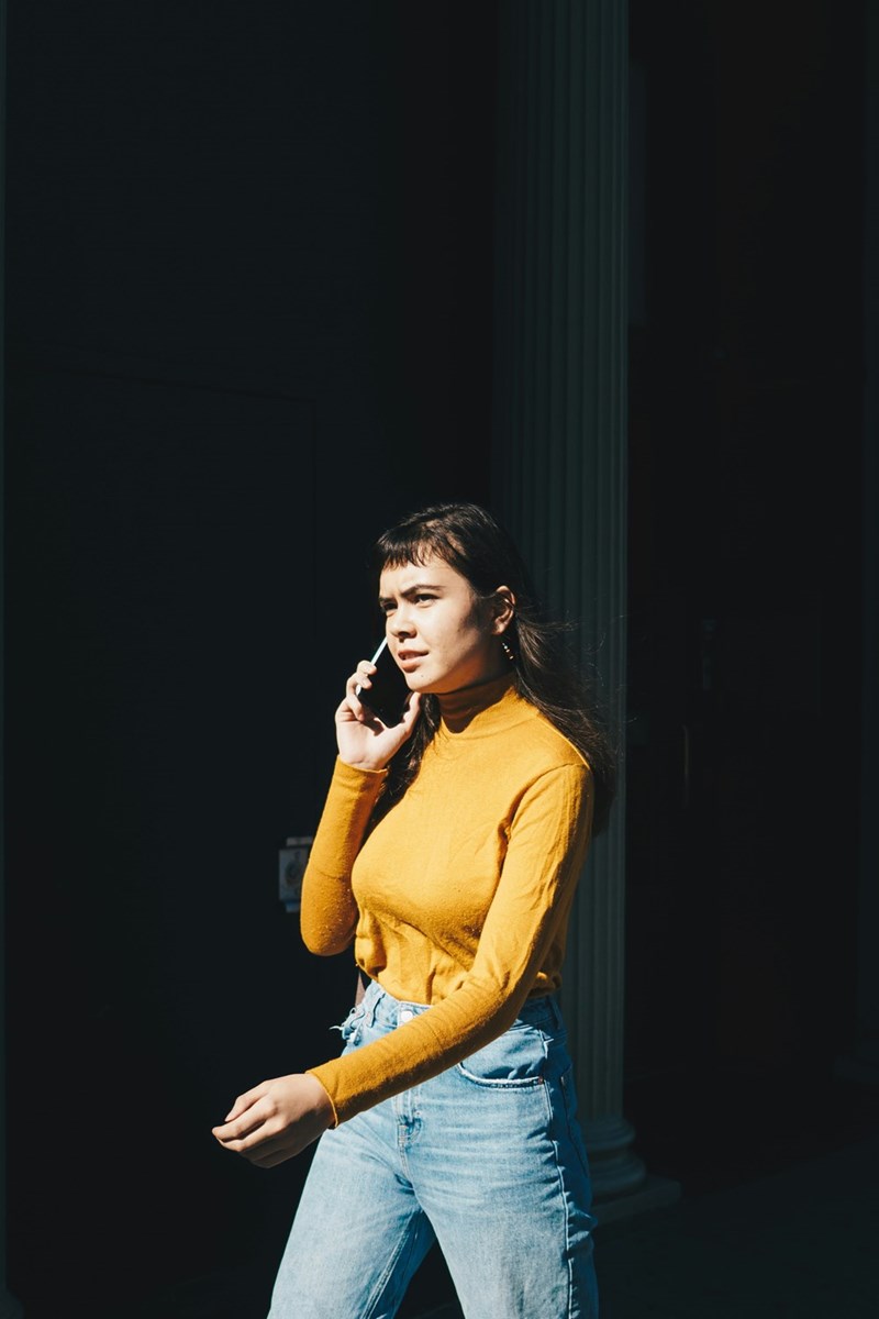 Woman in yellow top walking outdoors while talking on smartphone in sunlight.