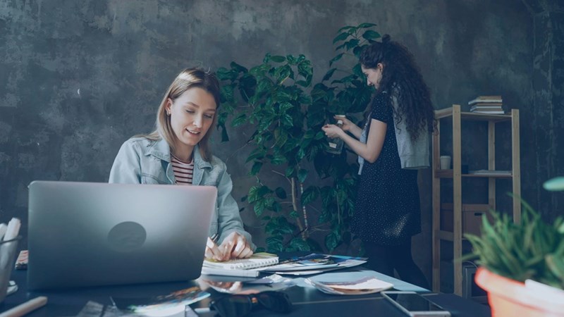 Woman using laptop at desk with papers while coworker waters plant in modern office workspace.