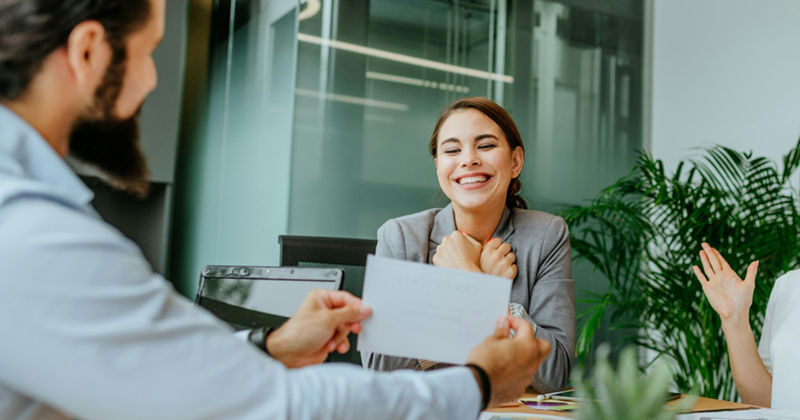A female employee crosses her arms across her chest as a male colleague presents a certificate to her across a meeting table