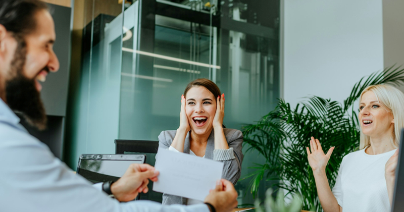 Two female colleagues look surprised as a male colleague presents a certificate across a meeting table