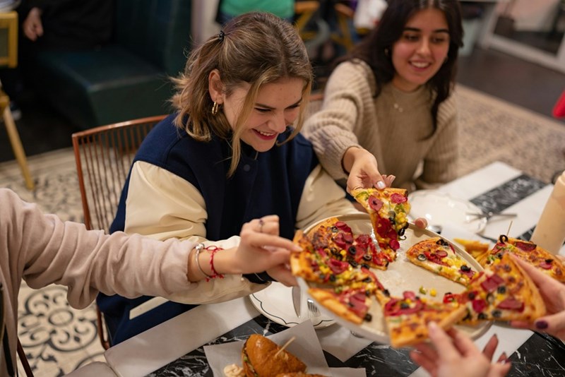 Group of teens grab slices of pizza while dining at a restaurant together.