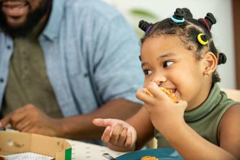 Little girl eats pizza slice while parent sits next to her.