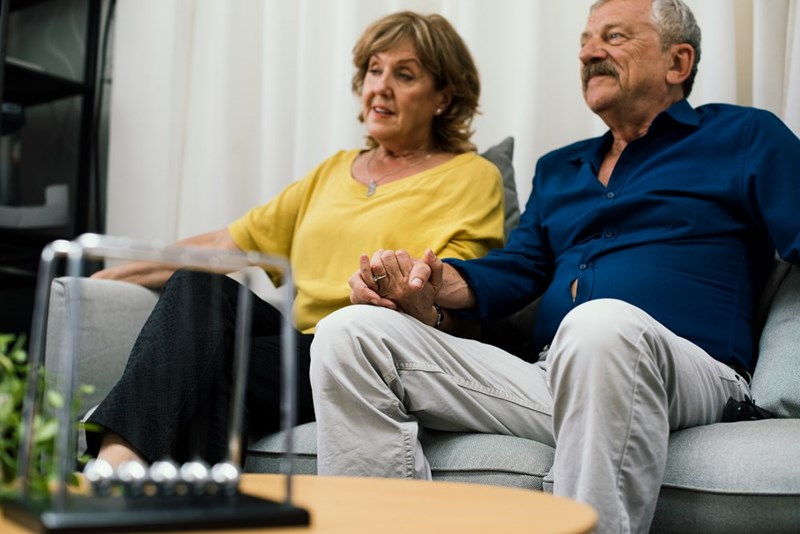 Old couple dressed casually sitting down on a grey couch holding hands.