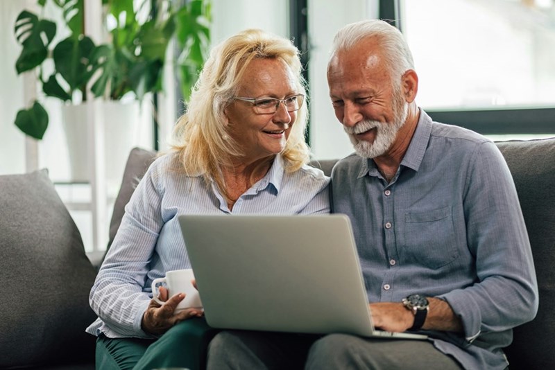 An old couple sitting down in a couch smiling and looking at a computer.
