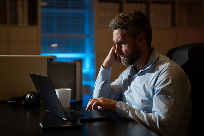 Man using laptop at night at desk with coffee, focused on work in low light office.