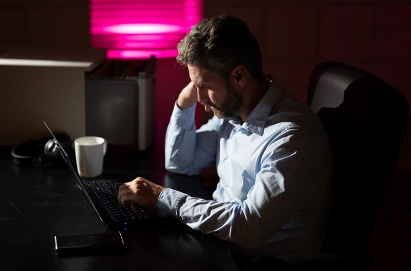 Man using laptop at night with neon light, focused on work at desk with coffee.