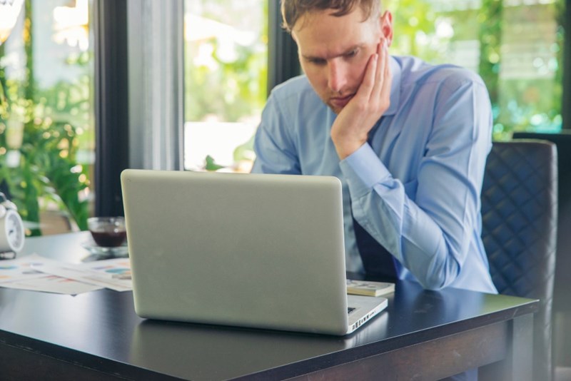 Stressed man working on laptop at desk, resting head on hand while reviewing documents at office.