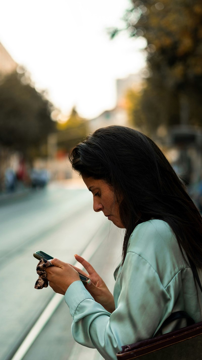 Woman checking smartphone on city street, looking concerned while standing near tram tracks at sunset.