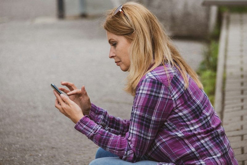 Woman in plaid shirt sitting outside, focused on her smartphone while typing a message on a quiet street.