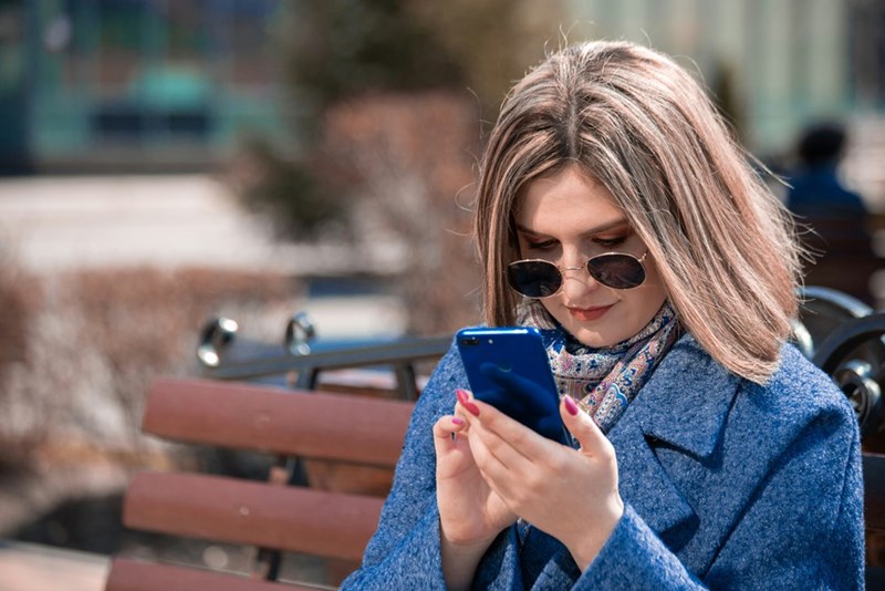 Woman in blue coat and sunglasses using smartphone while sitting on a park bench on a sunny day.