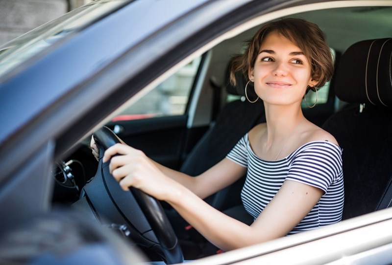 Smiling woman driving car, looking to the side with hands on steering wheel in daylight.